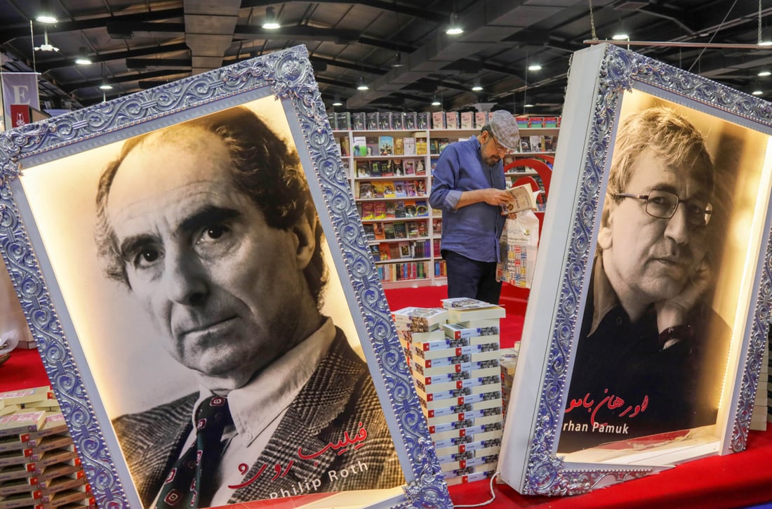 A visitor checks a book behind framed portraits of US novelist Philip Milton Roth (L) and Turkish writer Ferit Ohan Pamuk during the 17th Arbil International Book Fair, in the capital of the autonomous Kurdish region of northern Iraq, on April 9, 2025. (Photo by Safin HAMID / AFP)